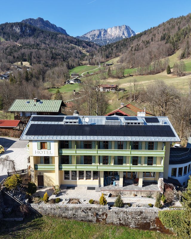Luftaufnahme eines Gebäudes mit Photovoltaikanlage auf dem Dach vor Bergen und ländlicher Umgebung.