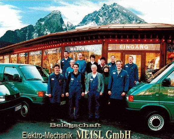 Belegschaft von Elektro-Mechanik MEISL GmbH vor Firmengebäude und Servicefahrzeugen, dahinter Berge.