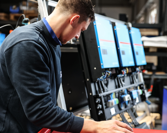Techniker arbeitet an einer Anlage mit mehreren Monitoren und verkabelten elektronischen Komponenten in einer Werkstatt.