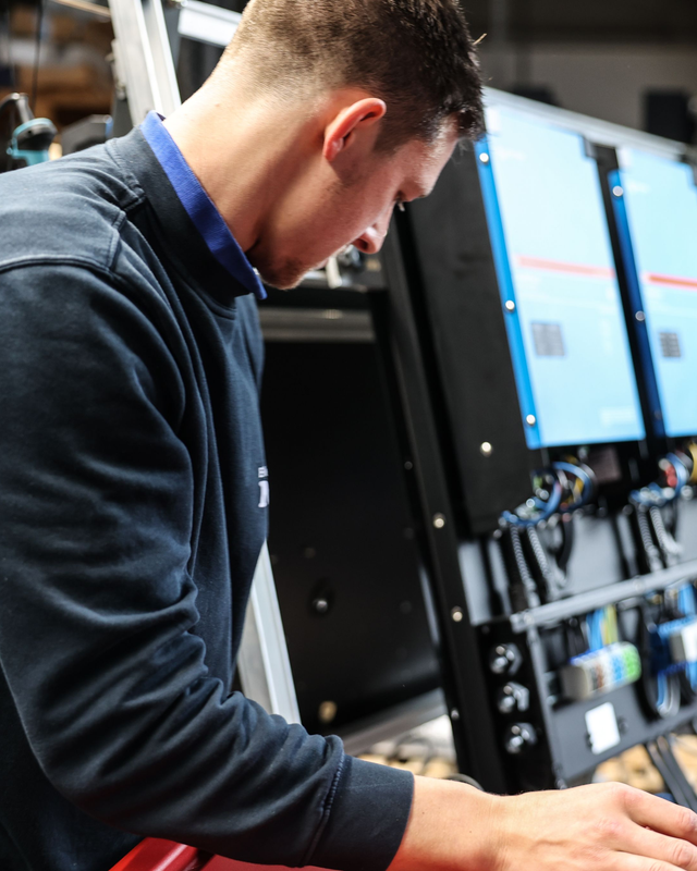 Techniker arbeitet an einer Anlage mit mehreren Monitoren und verkabelten elektronischen Komponenten in einer Werkstatt.