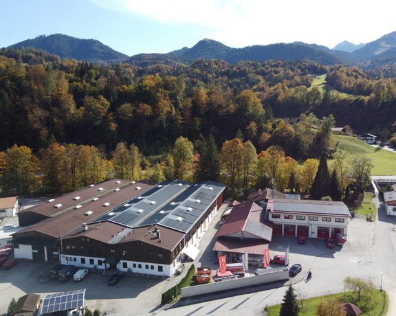 Luftaufnahme eines Firmengebäudes mit Solaranlage auf dem Dach vor Wald und Bergen.