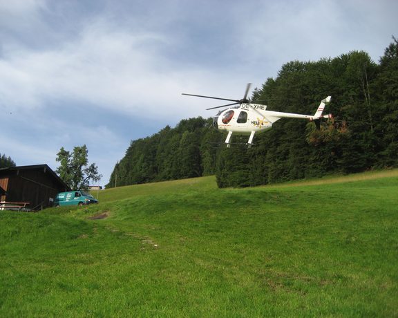 Ein Hubschrauber schwebt über einer Wiese am Waldrand neben einem Gebäude und einem Servicefahrzeug.