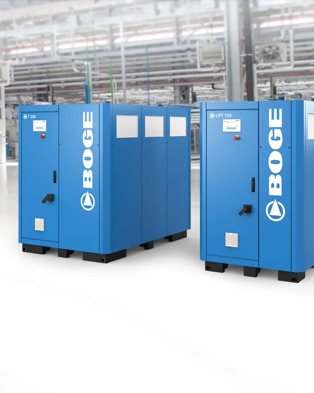 Zwei blaue BOGE-Druckluftkompressoren stehen in einer Industriehalle mit Rohrleitungen im Hintergrund.
