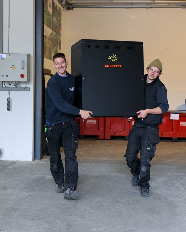 Zwei Mitarbeiter tragen einen schwarzen Energiespeicher in einer Lagerhalle.