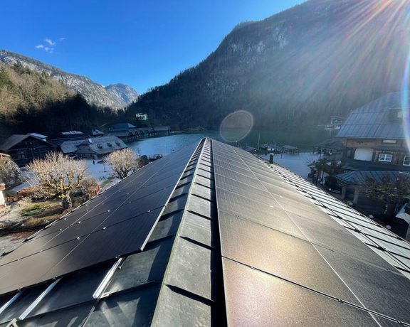 Photovoltaikmodule auf einem Hausdach in winterlicher Berglandschaft mit See und Gebäuden.