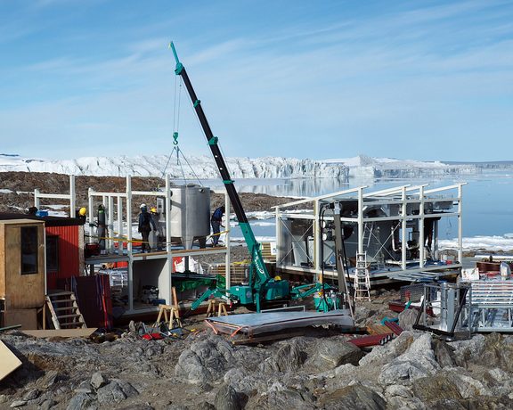 Montage einer Energieanlage mit Kran und Technikmodulen an einer felsigen Küste vor Eis und Wasser.